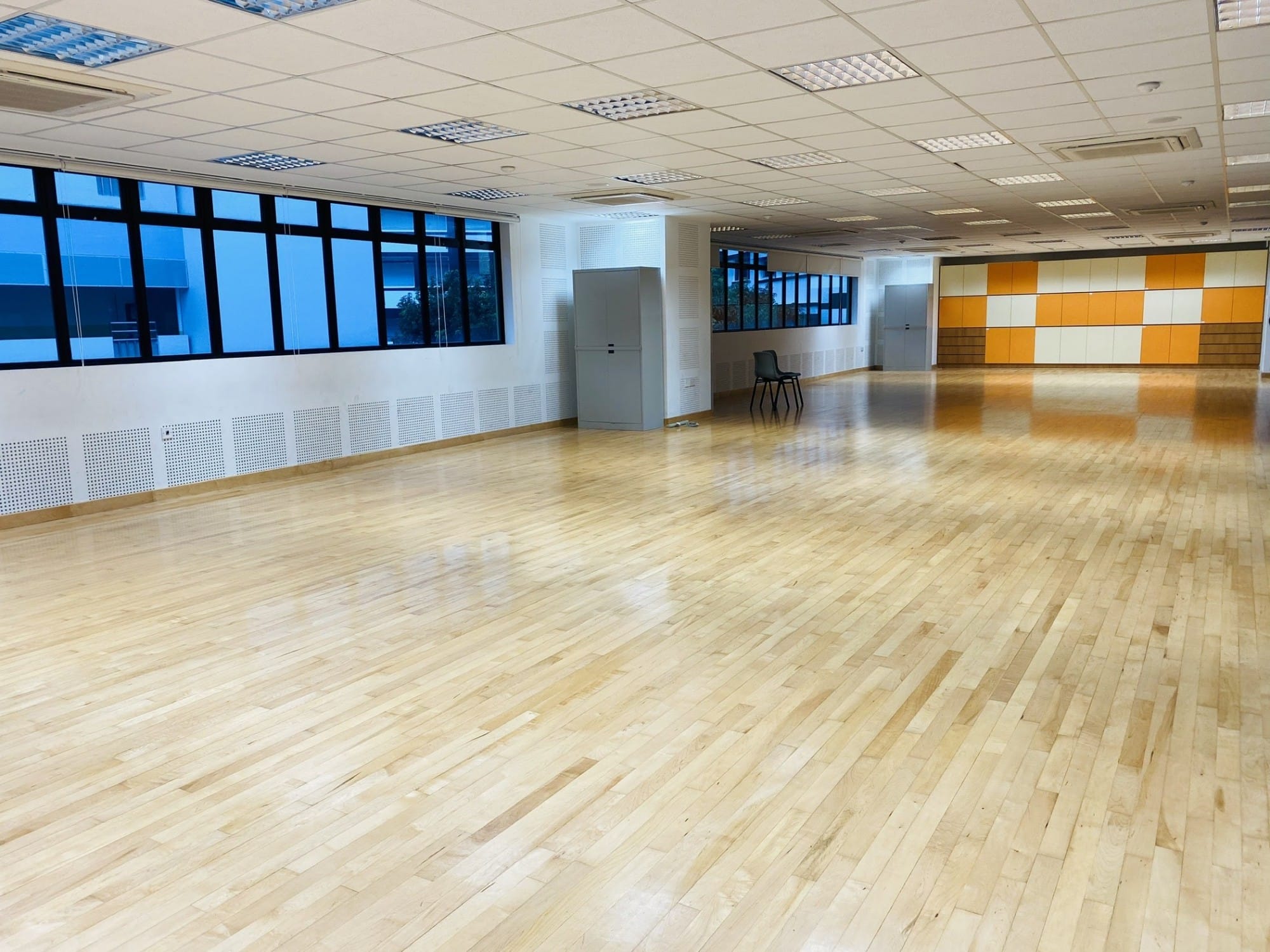 Large empty indoor hall with polished wooden floor, large windows, and an orange and white checkered wall panel.